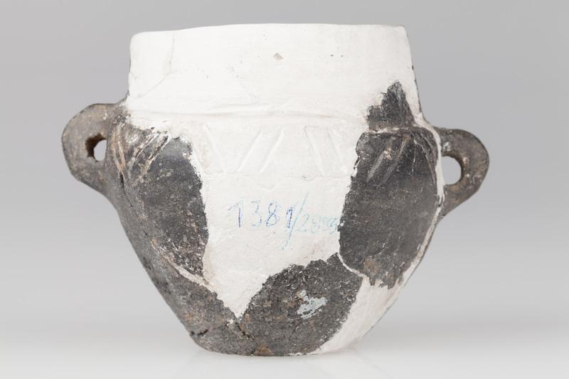 Waza czerniona z wysoką szyjką, baniastym brzuścem i płaskim dnem. Naczynie ornamentowane dookolną linią rytą.