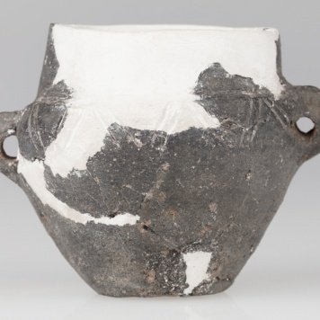 Waza czerniona z wysoką szyjką, baniastym brzuścem i płaskim dnem. Naczynie ornamentowane dookolną linią rytą.